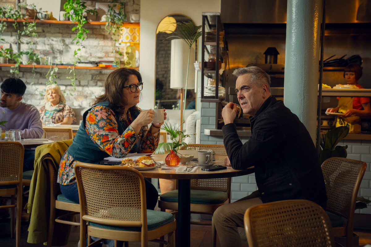 Two people sit at a cozy restaurant table engaged in conversation, surrounded by plants and warm lighting. The atmosphere is relaxed and intimate, with a chef visible in the open kitchen behind them.
