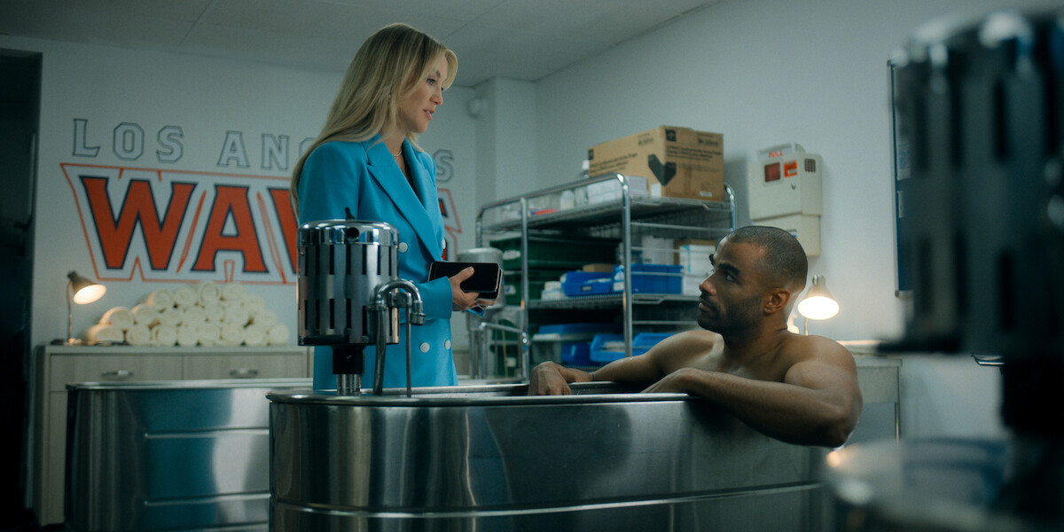 A woman in a blue coat talks to a man sitting in an ice bath in a locker room with towels, shelves, and Los Angeles Waves signage on the wall.