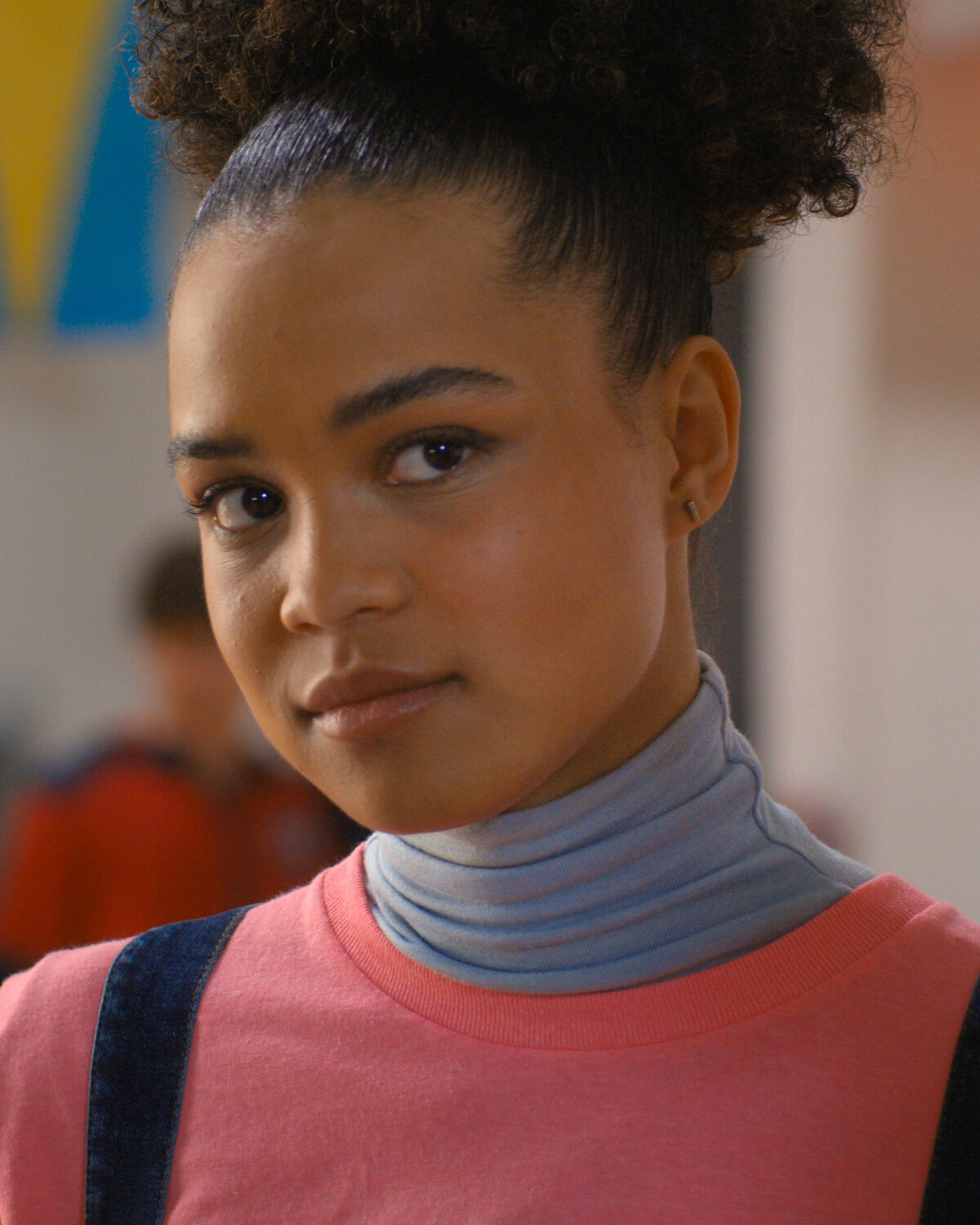 Teen girl with curly hair pulled up, wearing a pink shirt over a light blue turtleneck and denim overalls, standing indoors with a soft, neutral background and colorful banners.
