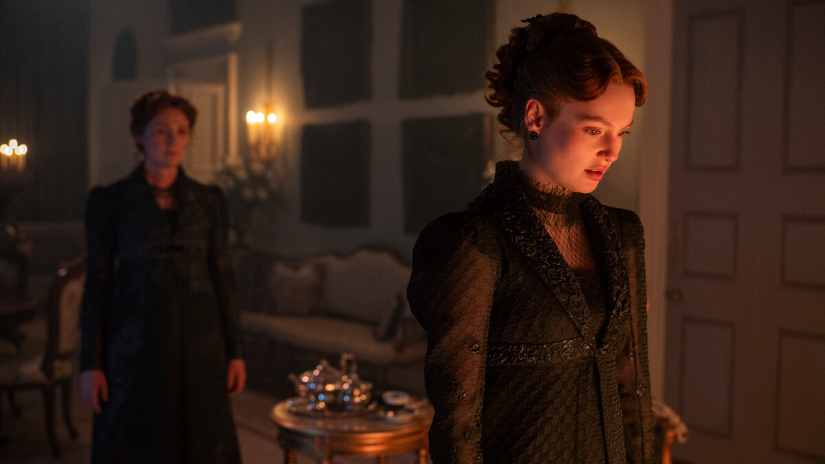 Two women in dark Victorian dresses stand in a dimly lit, elegant room with classical furniture, a tea set on a small table, and candles glowing softly in the background. The atmosphere appears tense and dramatic.