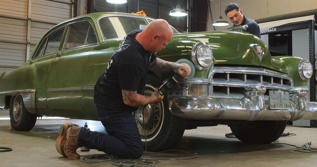 Two men in an auto body shop work on a vintage green vehicle.