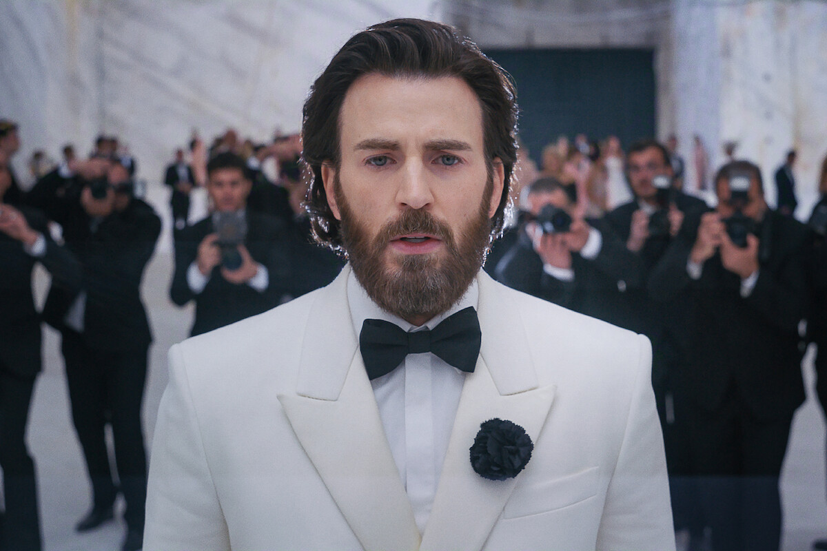 A man in a white tuxedo with a black bow tie stands in a marble hall, facing the camera, while photographers in black suits take pictures in the background. Formal, elegant setting.