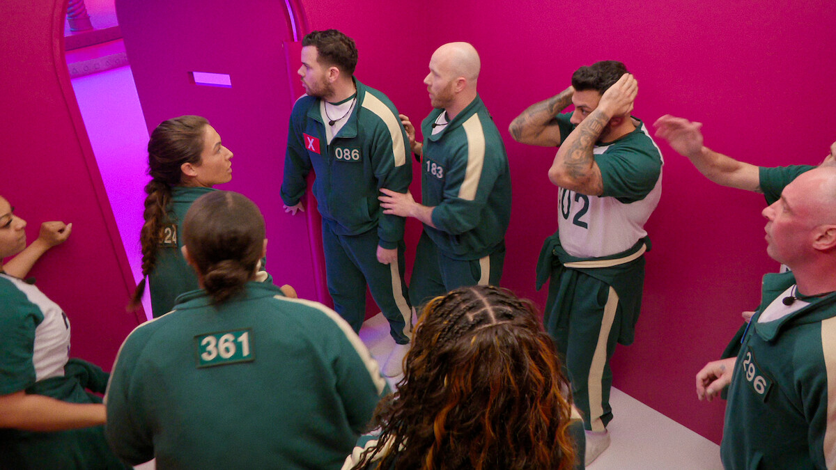 A group of people in green uniforms with numbers stand in a brightly lit, pink-walled hallway, appearing tense and alert as they look toward an open door, creating a mood of suspense and anticipation.