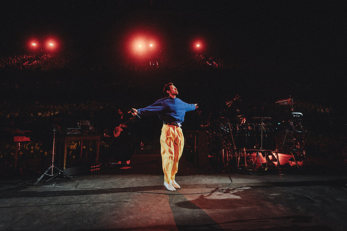 Harry Styles in blue top and yellow pants jumps with arms outstretched on a dimly lit concert stage, surrounded by musical instruments, with red lights and a large audience in the background.