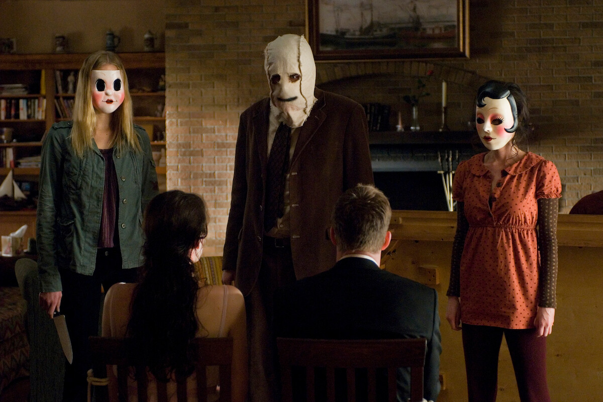 Three masked figures stand threateningly in a dimly lit living room facing two seated people with their backs to the camera, creating a tense and ominous atmosphere.
