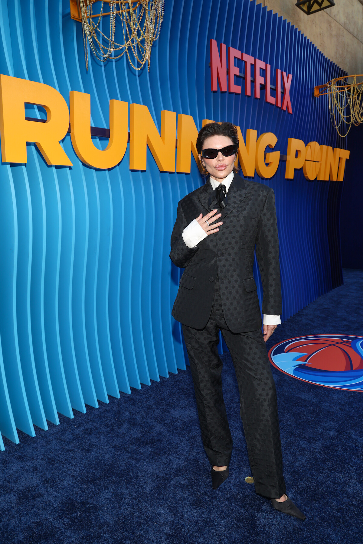Person in a dark patterned suit and sunglasses poses on a blue carpet in front of a large Netflix "Running Point" promotional backdrop with bold yellow lettering and decorative lighting fixtures, creating a formal event atmosphere.