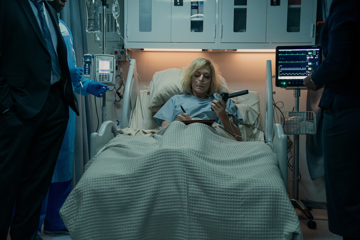 Woman in a hospital bed wearing a gown, surrounded by medical equipment and monitors, holds a notepad and pen. Two people stand nearby, partially visible, in a dimly lit hospital room setting.