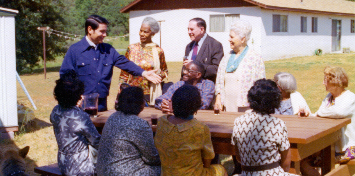 A group of people, diverse in age and ethnicity, gather outdoors around a wooden picnic table on a sunny day, appearing cheerful and engaged in conversation with a house and green lawn in the background.