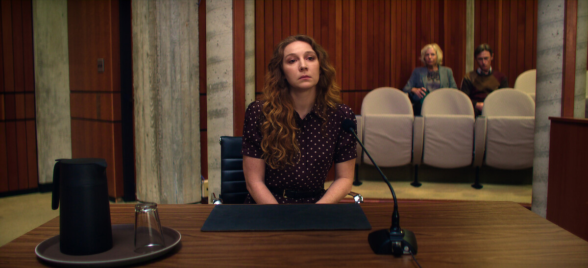 A woman with long hair sits at a witness stand in a courtroom, facing forward. There is a microphone, a carafe, and a glass on the table. Two people are seated in the gallery behind her, in front of wood paneled walls.