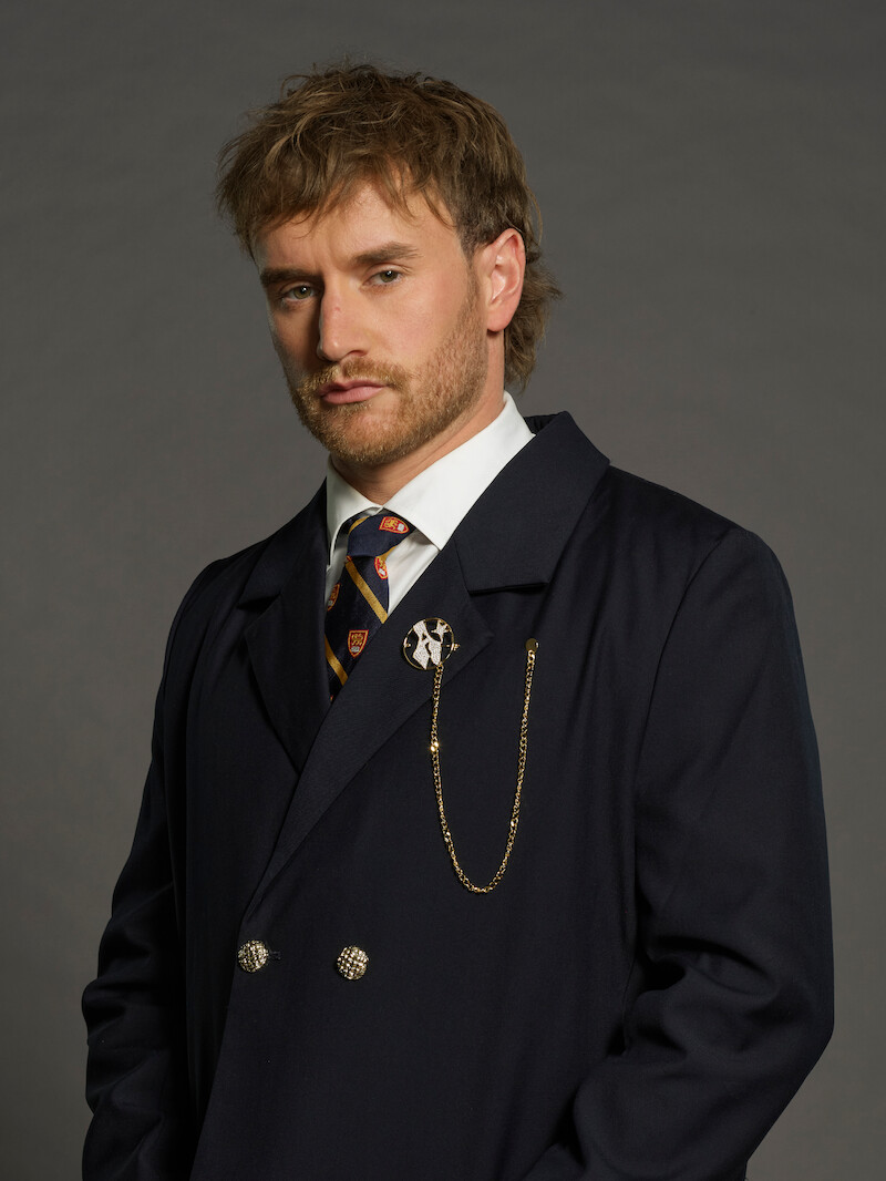 Homem com cabelo e barba castanho-claros posa em frente a um plano de fundo neutro, usando um terno azul-marinho com uma gravata e uma corrente dourada, com uma expressão séria no rosto.