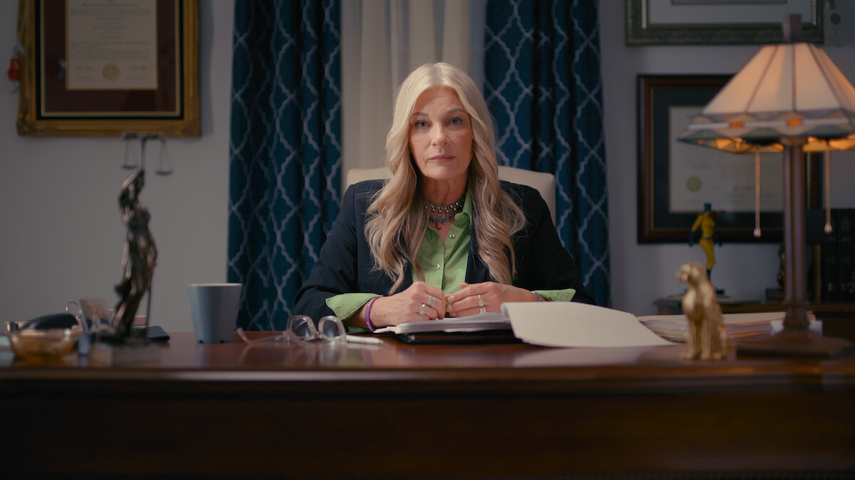 A woman with long blonde hair sits at an office desk with papers, a lamp, and legal decorations, in front of blue curtains and framed certificates, appearing professional and attentive.