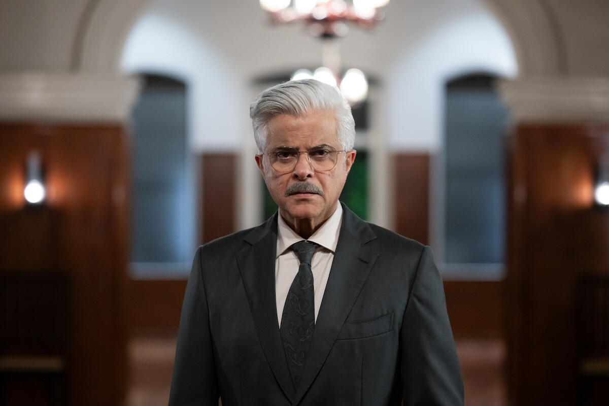 Older man in a dark suit and tie standing in a formal, well-lit hallway with arched doorways and wood-paneled walls, looking serious and focused.