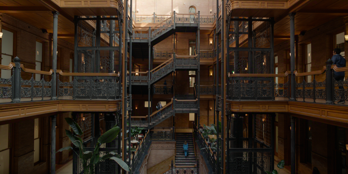 Victorian-style multi-story building interior with ornate iron railings, open walkways, central staircase, exposed brick walls, natural light, potted plants, and a few people walking or standing on different levels.