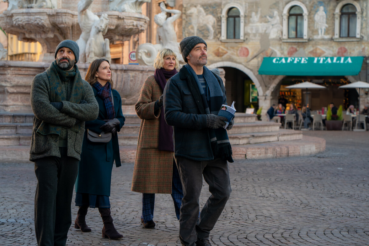 Four adults in winter clothing walk through a European plaza with a stone fountain and historic buildings, including a café with outdoor seating, in the background.