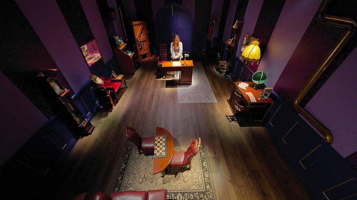 Mulher em pé diante de uma escrivaninha em uma sala elegante e vintage com paredes roxas, piso de madeira, mesa de xadrez, cadeiras de couro, estantes de livros, abajures e decoração aconchegante, vista de cima.