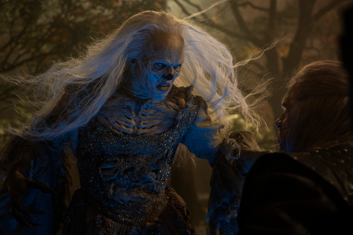 An undead creature with long white hair and decayed skin confronts a person in dark clothing in a misty, eerie forest setting at night, creating a tense, supernatural atmosphere.