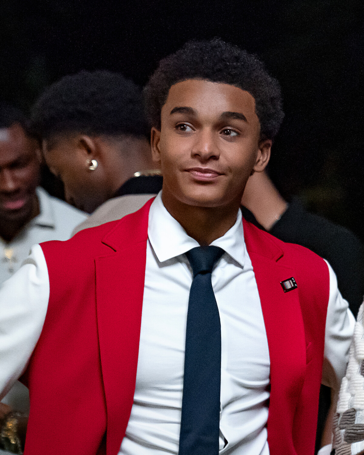 Young man in formal attire with a red blazer, white shirt, and dark tie standing at an indoor event. Other people in formalwear are in the background, the setting appears social and lively.