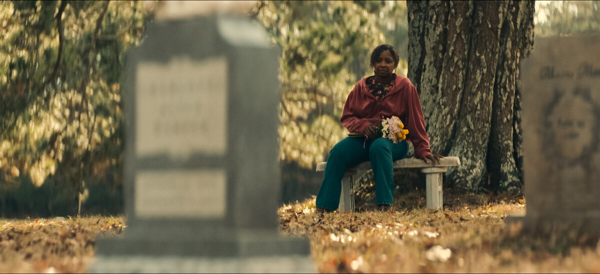A woman sits alone on a bench in a cemetery holding flowers, surrounded by gravestones and trees.