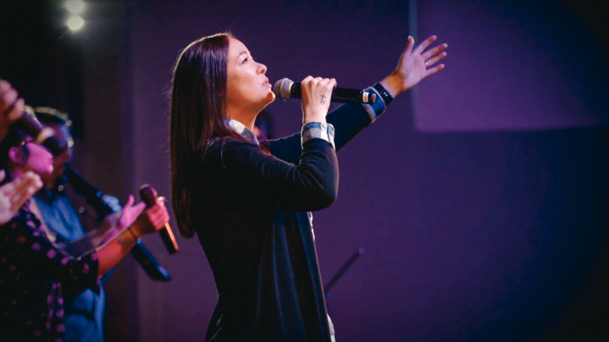 Woman singing passionately into a microphone on stage, with colorful stage lighting and other musicians visible in the background, suggesting a live music or worship performance setting.