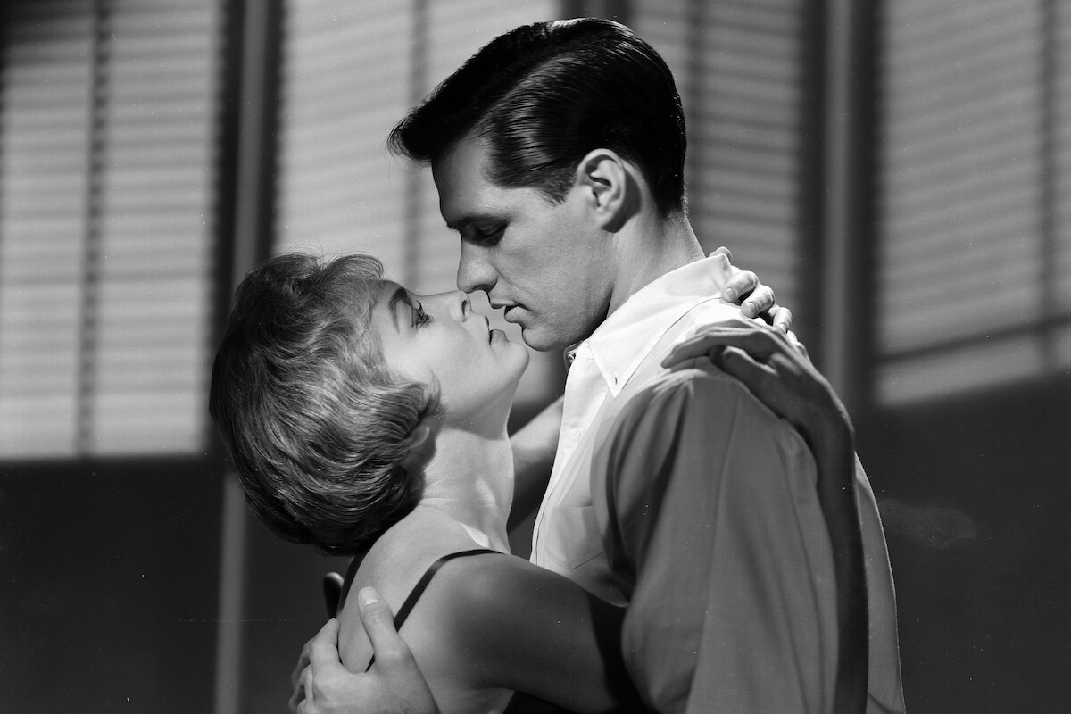 A man and woman embrace and prepare to kiss in a dramatic black and white scene, standing indoors with window blinds in the background.