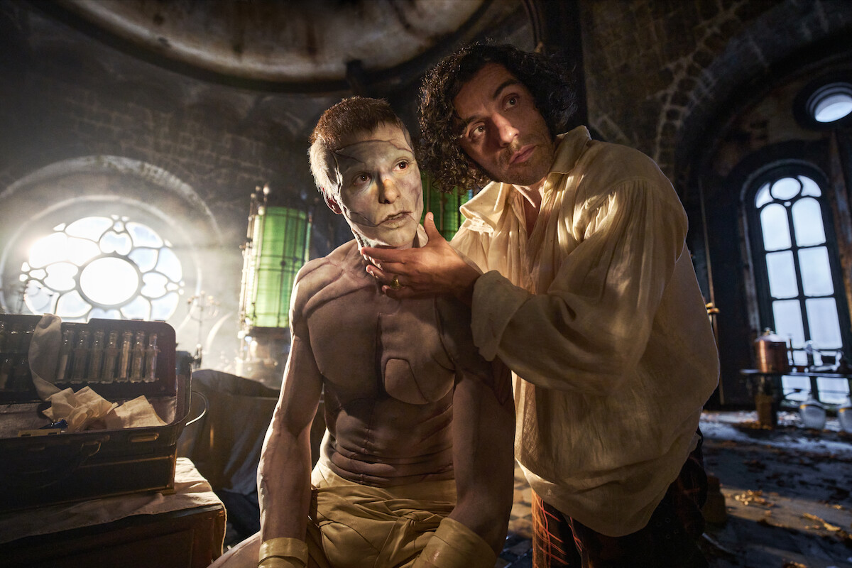 A man applies white makeup to a shirtless figure in a dim, gothic laboratory bathed in dramatic light, evoking a mysterious, tense, and atmospheric mood with vintage scientific equipment in the background.