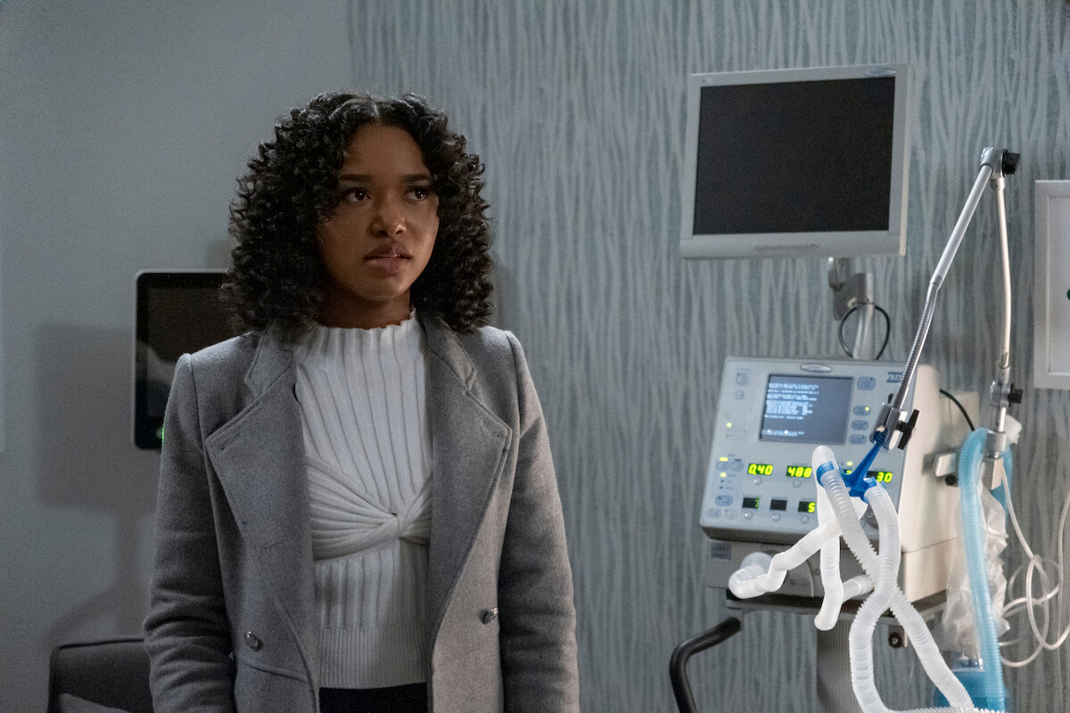 Bailey Tippen as Sylvie stands in a modern hospital room next to medical equipment and monitors, wearing a gray coat and white sweater, looking concerned.