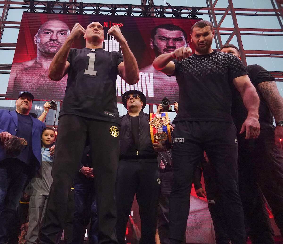 Two fighters pose on stage at a weigh-in event, surrounded by their teams and media, with a large promotional fight banner featuring their faces and names displayed in the background inside a modern, well-lit venue.