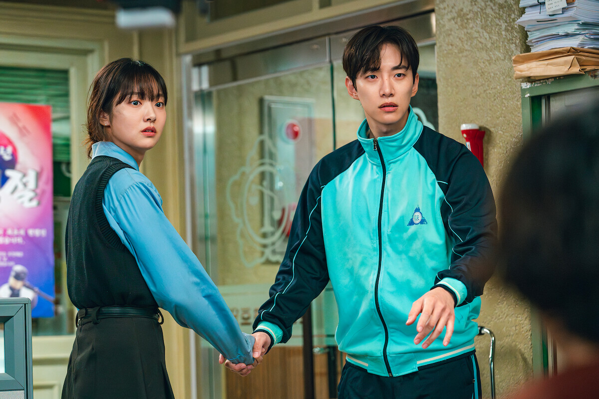 A woman and man hold hands, looking surprised or alarmed, inside a cluttered office with paperwork and posters on the wall, under fluorescent lighting.