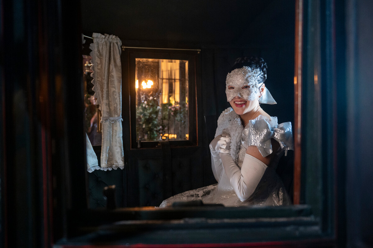 Em uma carruagem à meia-luz, com cortinas rebuscadas e uma janela iluminada ao fundo, uma mulher de máscara e vestido branco elegante sorri, sentada, em um clima misterioso e festivo.