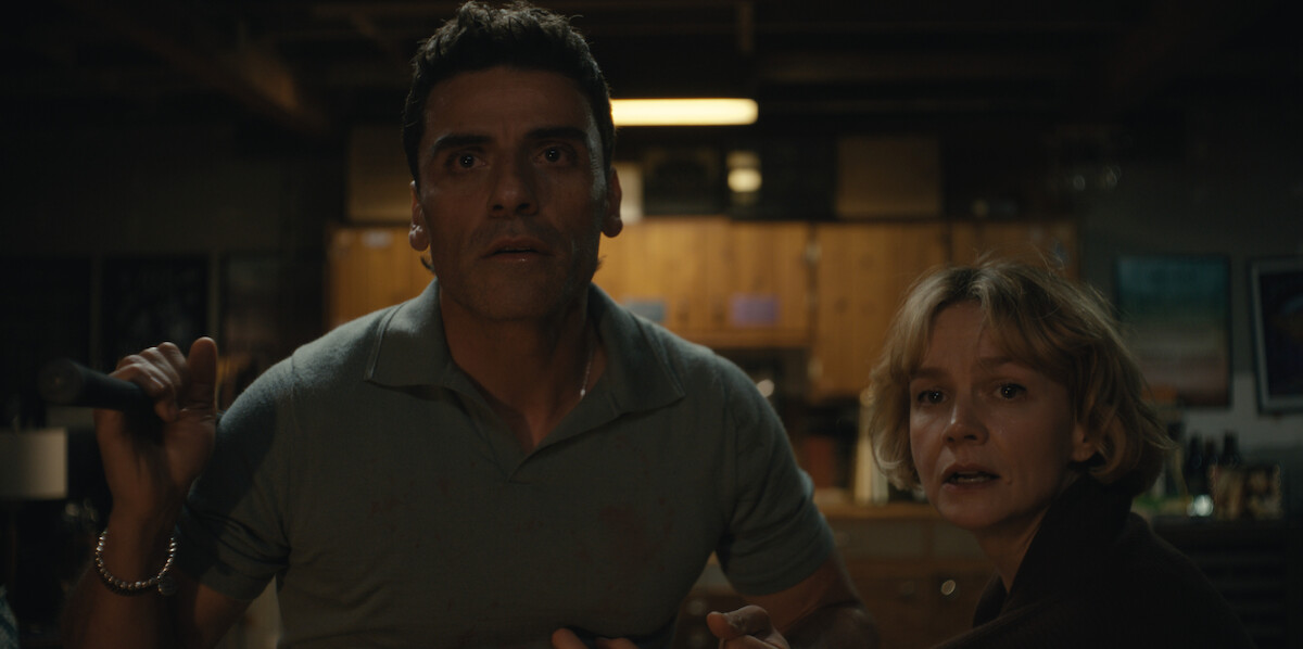 A man and woman in a dimly lit, cluttered kitchen basement look intensely forward, appearing tense and alert.