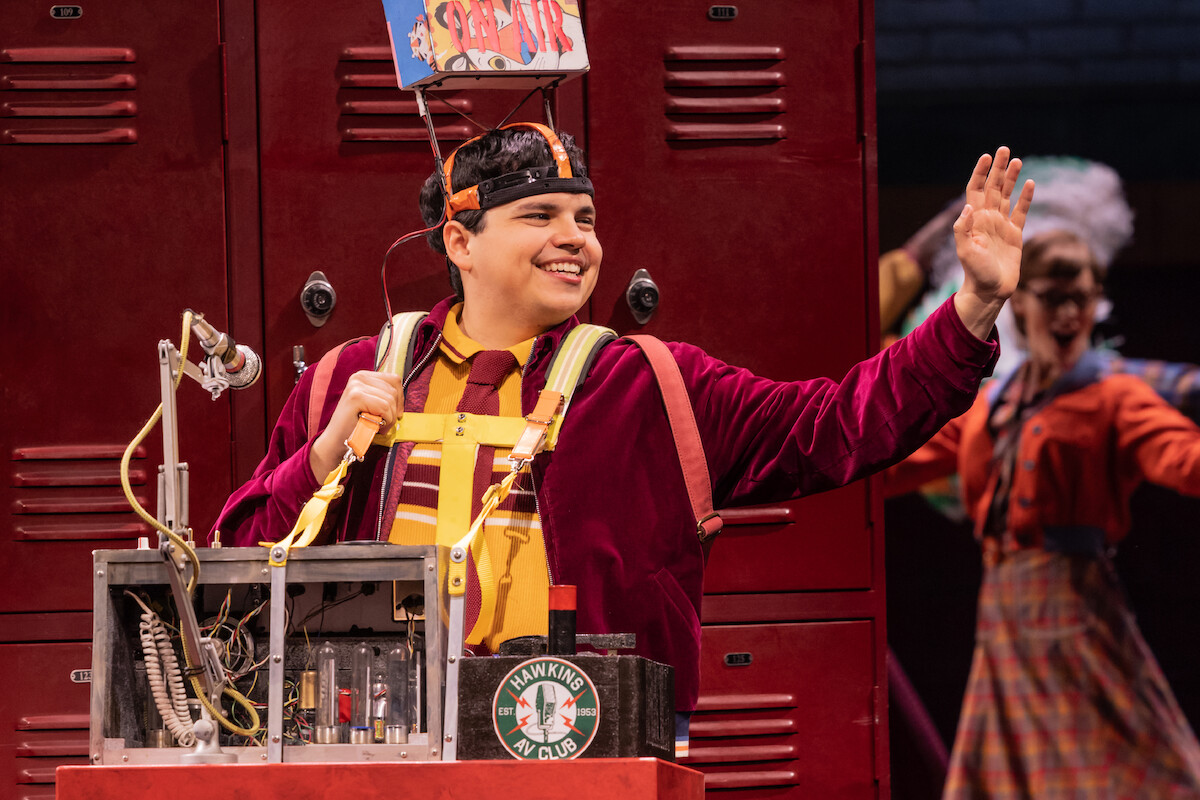 Smiling actor in colorful costume with headgear and mechanical backpack stands in front of red lockers on stage, waving, with another performer and theater props in the background.