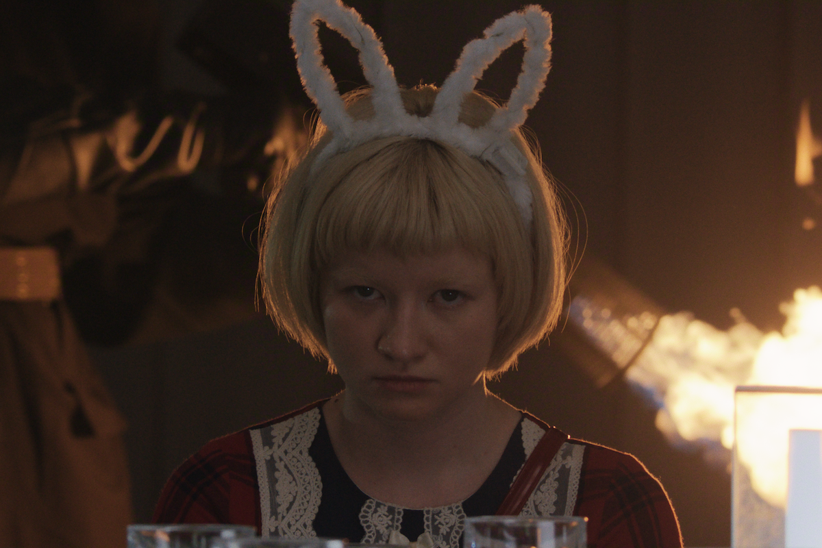 A serious-looking person with blond hair wearing white bunny ears sits indoors, with glasses on the table and fire burning in the background, suggesting a tense or dramatic scene.