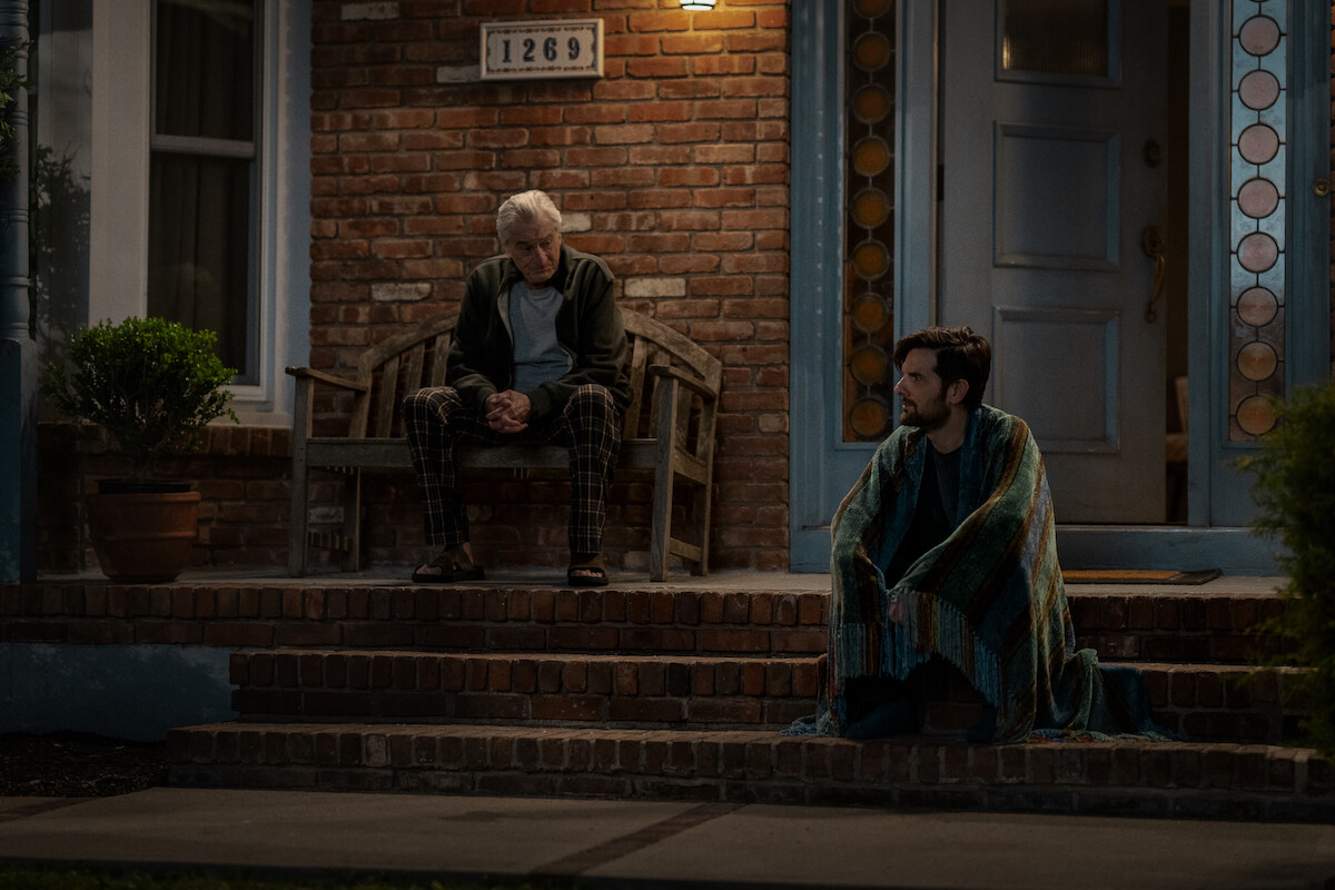 Two men sit outside a brick house at night; one on a bench looking down, the other wrapped in a blanket on the porch steps, suggesting a quiet, contemplative moment. The porch is dimly lit, evoking a calm, reflective mood.