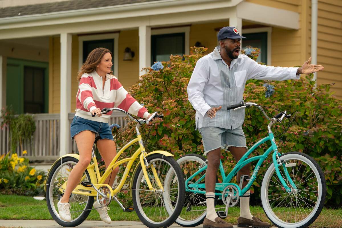 Two adults riding colorful bicycles in a suburban neighborhood, passing by a yellow house with a porch and garden, engaging in conversation outdoors on a sunny day.