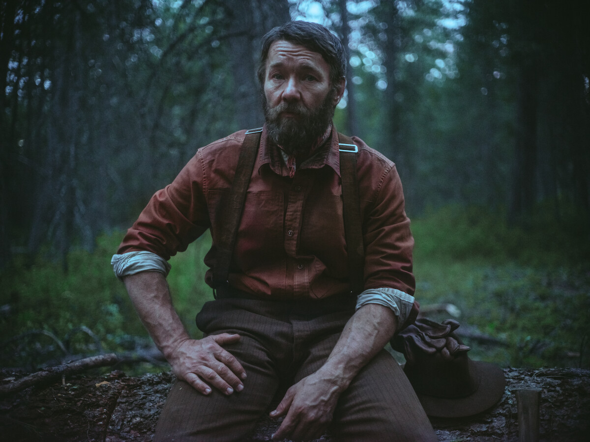 Bearded man in rugged clothing sits on a log in a dense, dimly lit forest, holding his hat beside him, looking worn and contemplative; the mood is somber and reflective in a wilderness setting.