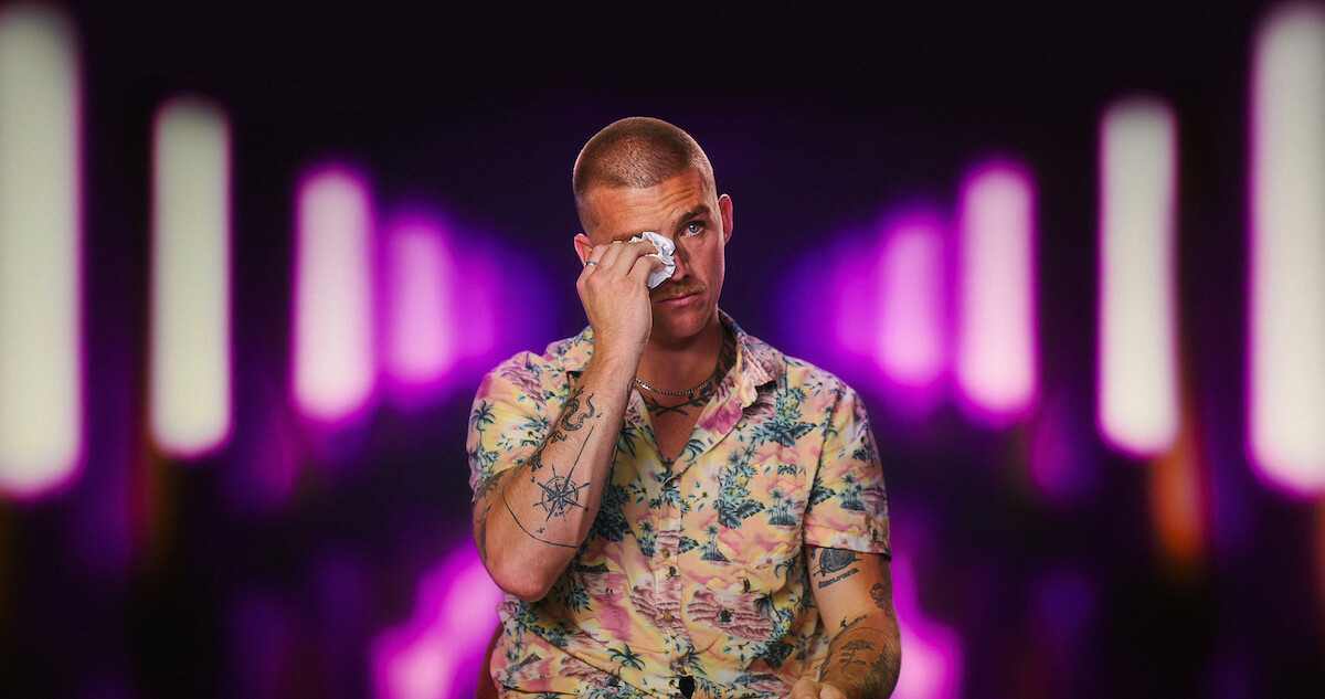 A man in a floral shirt wipes his eye, sitting in front of a blurred background with vertical neon purple and white lights, giving a dramatic nightclub or studio environment.