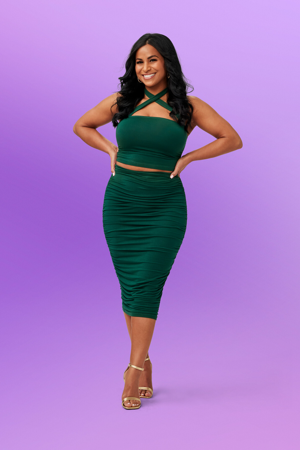 Brittany W. in a dark green dress and heels poses, smiling with hands on hips, against a plain light purple gradient studio background.