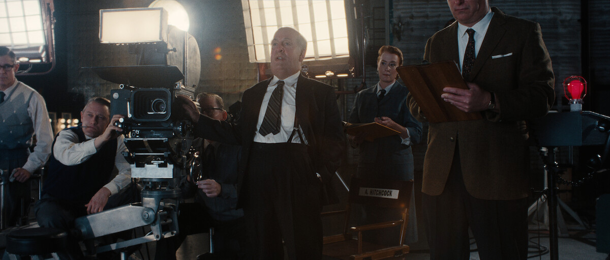 A man in a suit stands beside a large film camera on a movie set, surrounded by crew members and bright studio lights.