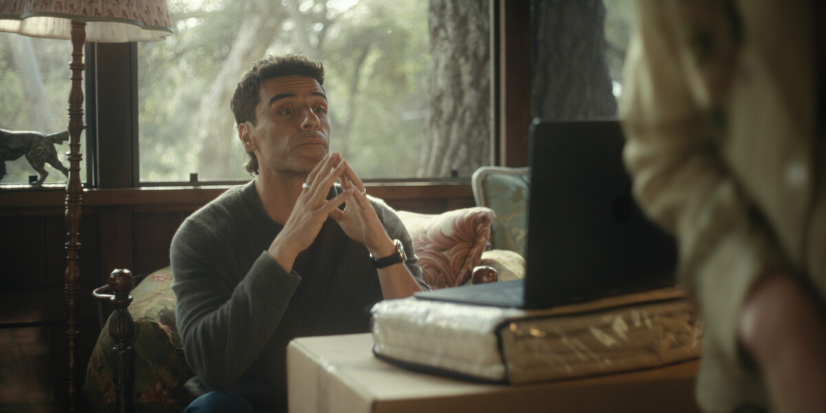 A man sitting indoors on a floral chair by a window, hands clasped near his face, looking at someone off-frame. A desk with a laptop and large book or binder is in front of him, and trees are visible outside.
