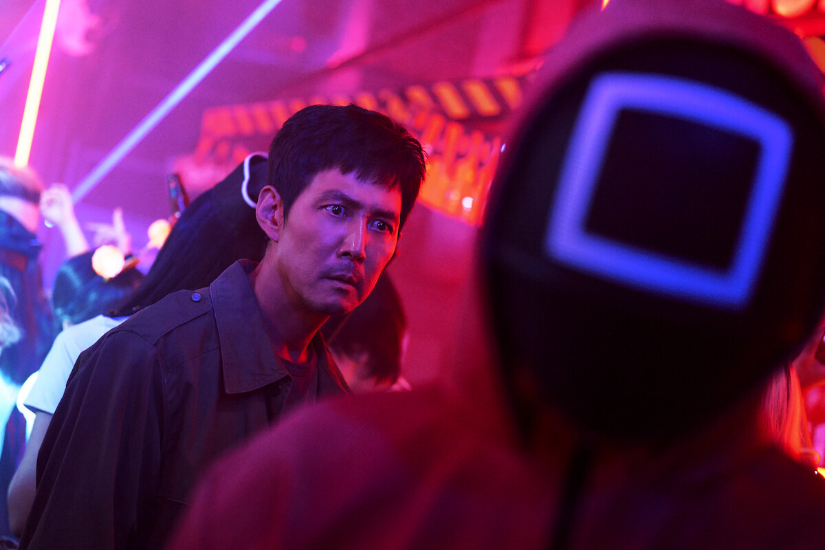 Lee Jung-jae with a tense expression in a vibrant, neon-lit environment, facing a mysterious figure in a hooded red suit with a glowing square mask, surrounded by a crowd and glowing lights.