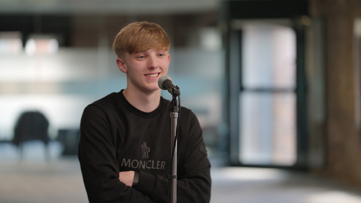 Young man in a Moncler sweatshirt stands smiling at a microphone in a modern, softly lit indoor space with glass doors and blurred furniture, suggesting a relaxed and casual atmosphere.