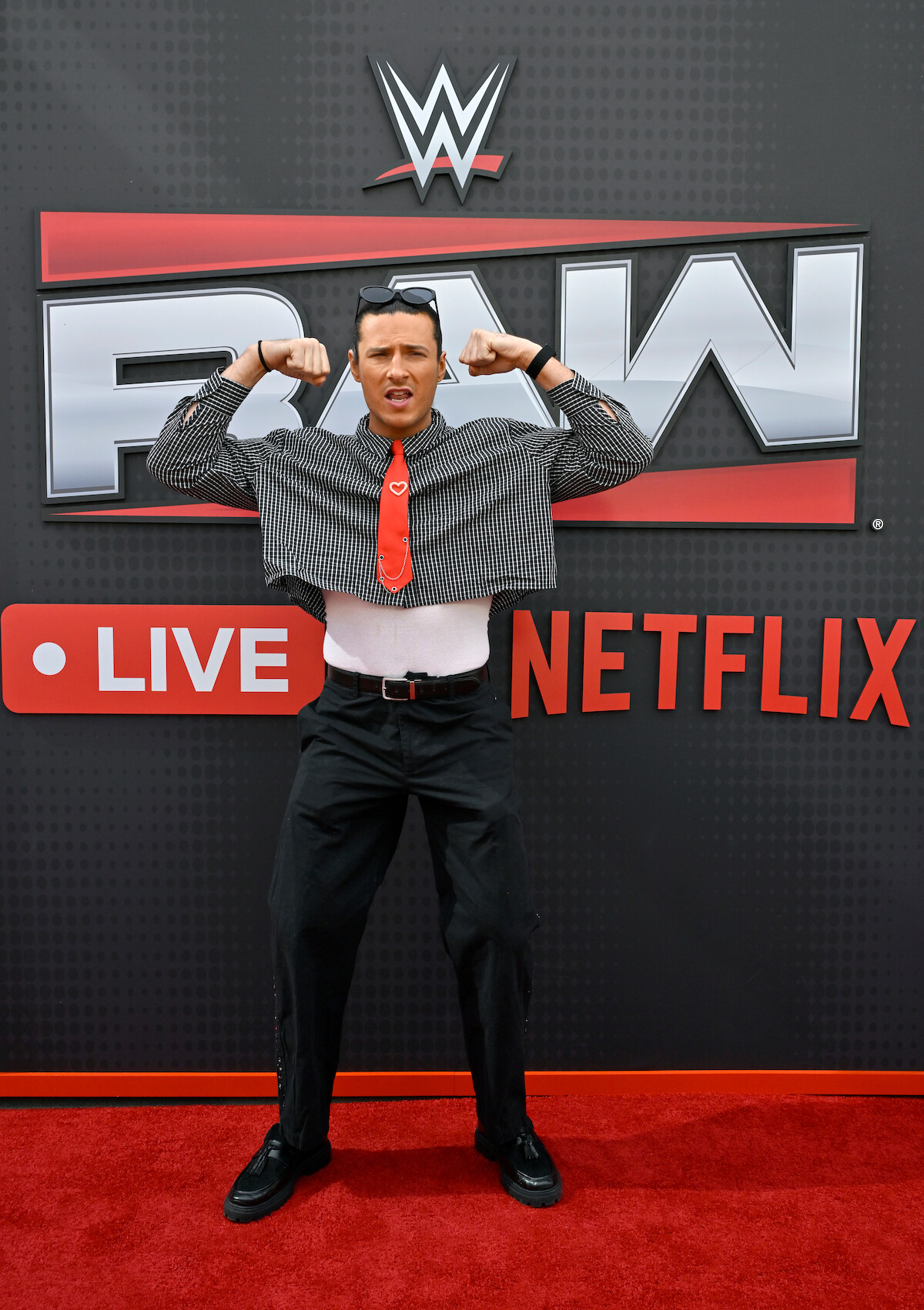 Person in quirky attire flexes in front of WWE RAW Live Netflix backdrop on a red carpet at a media or promotional event.