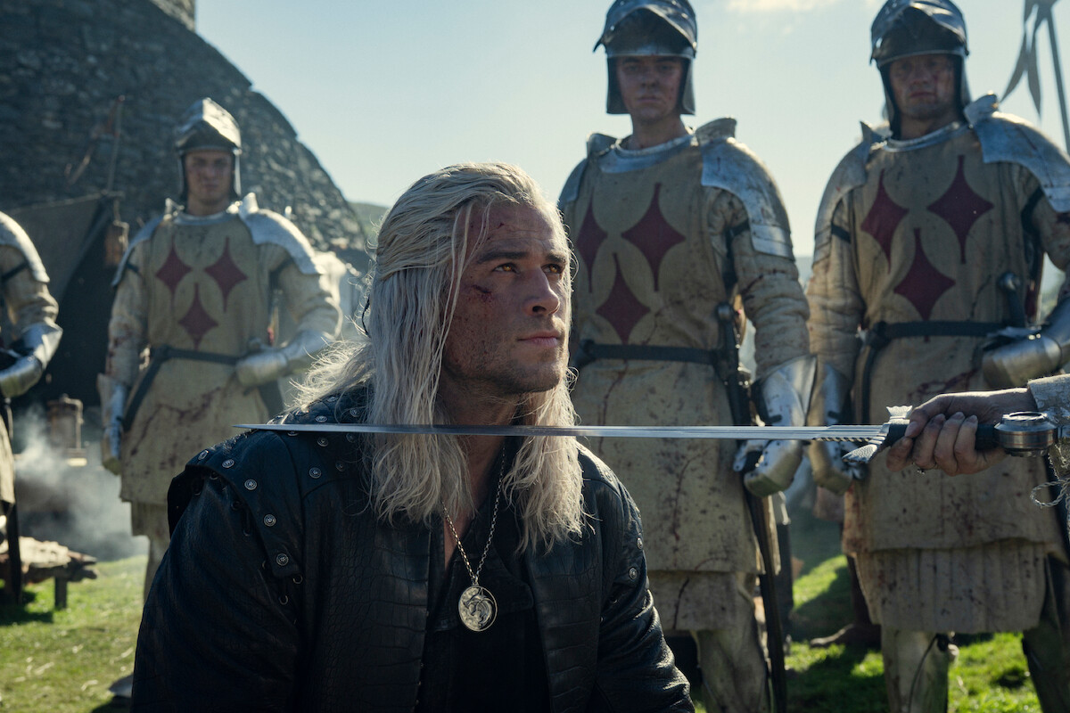 A man with long white hair kneels outdoors, sword at his neck, surrounded by armored soldiers with red emblems. The setting is medieval, tense, and dramatic, with strong sunlight casting shadows on the scene.