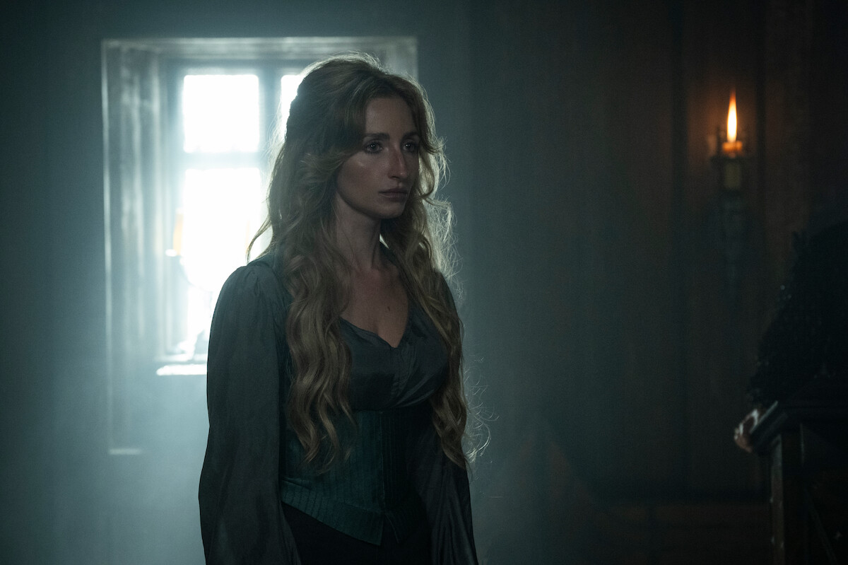 A solemn woman with long wavy hair stands in a dimly lit room with a glowing window behind her and a candle on the wall, creating a moody, dramatic atmosphere.