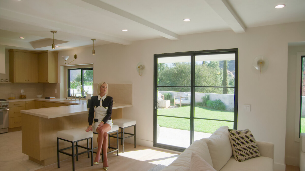 Woman sitting on a stool in a modern, sunlit kitchen and living room with large windows overlooking a green backyard, creating a bright and relaxed atmosphere.