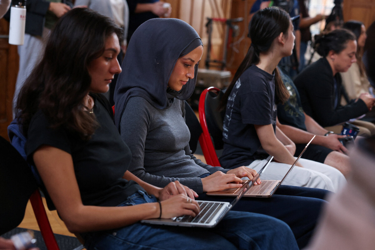 Women seated in rows at an indoor space, some using laptops, in a focused and attentive environment with wooden walls and a casual, collaborative setting.
