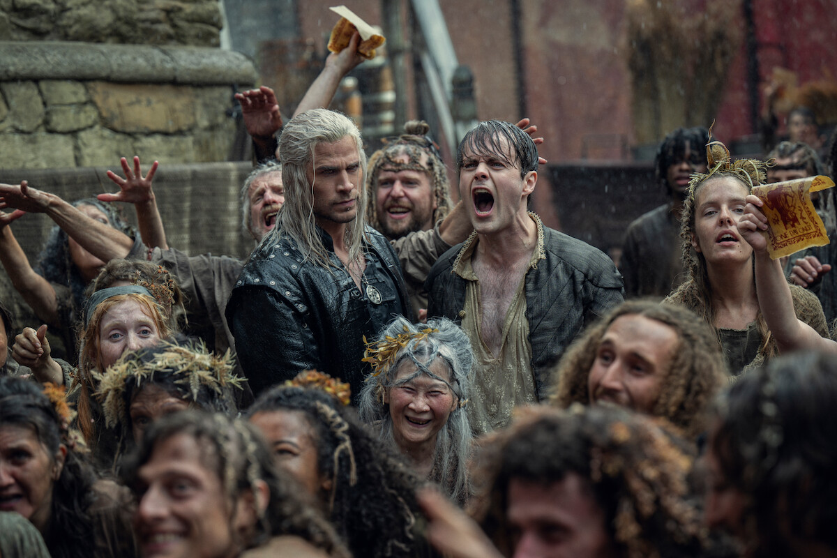 Two men, one with white hair and the other shouting, stand amid a lively, muddy medieval crowd celebrating outdoors. The mood is intense and chaotic, with expressions of joy and excitement.