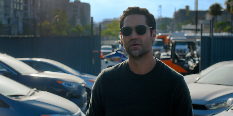 Man wearing sunglasses standing in a parking lot with several cars and city buildings in the background, daytime, urban setting, serious mood.