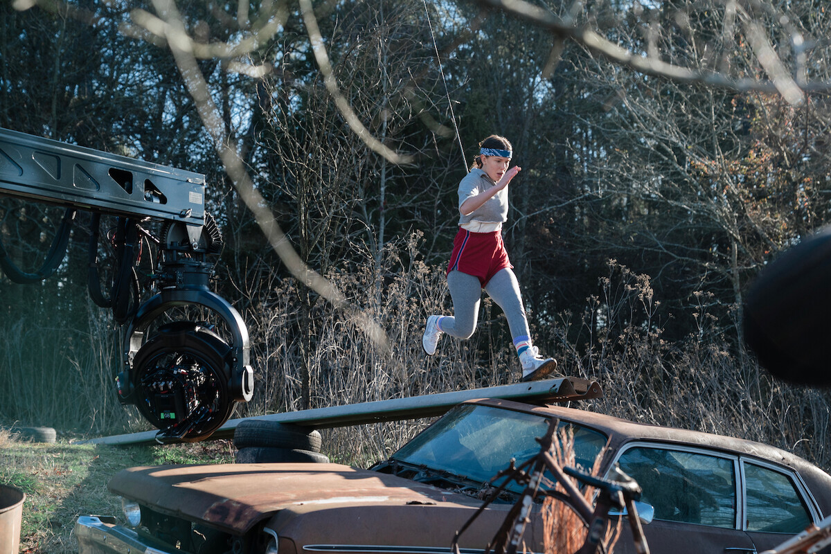 Young actor in athletic clothes sprints along a narrow plank above a rusty car as a camera rig tracks alongside.