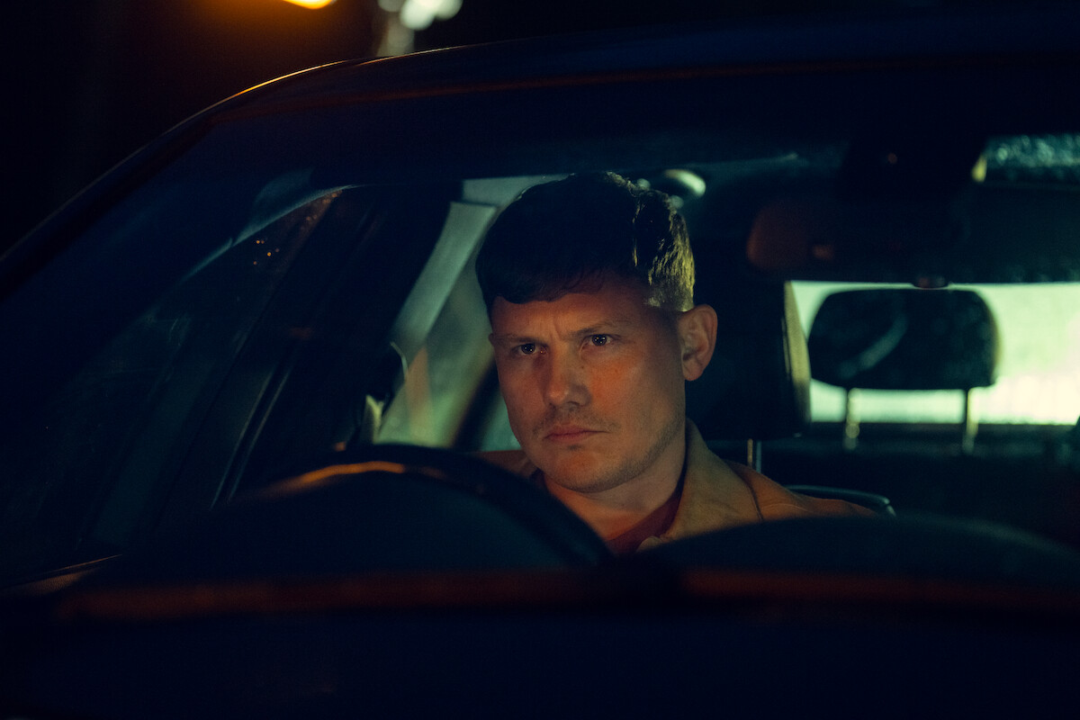 A serious-looking man sits alone in a car at night, illuminated by moody, dim lighting from outside, suggesting a tense or contemplative atmosphere.